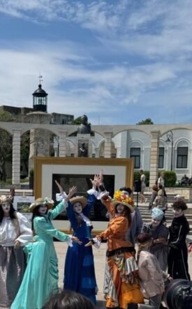 Group of performers in vintage costumes posing and dancing in a sunny outdoor plaza; audience watches in the background.