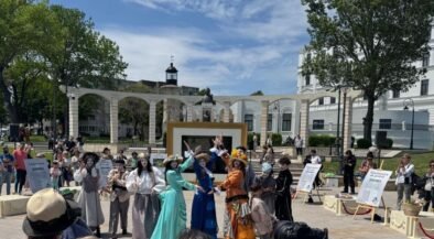 Group of performers in vintage costumes posing and dancing in a sunny outdoor plaza; audience watches in the background.