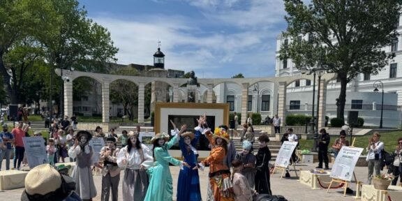 Group of performers in vintage costumes posing and dancing in a sunny outdoor plaza; audience watches in the background.
