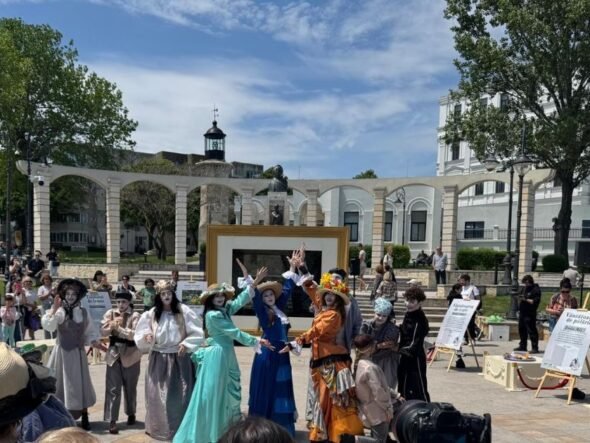Group of performers in vintage costumes posing and dancing in a sunny outdoor plaza; audience watches in the background.