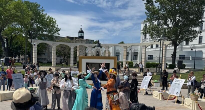 Group of performers in vintage costumes posing and dancing in a sunny outdoor plaza; audience watches in the background.