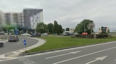 Roundabout with a grassy median, several cars, and a spiky spherical sculpture in the center; buildings in the background under a cloudy sky.
