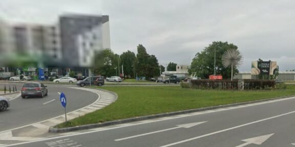 Roundabout with a grassy median, several cars, and a spiky spherical sculpture in the center; buildings in the background under a cloudy sky.