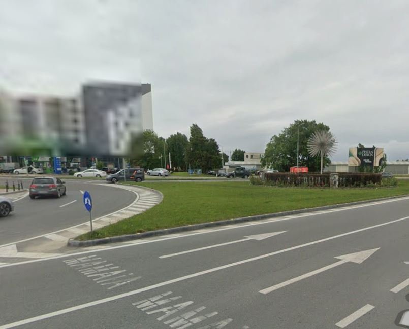 Roundabout with a grassy median, several cars, and a spiky spherical sculpture in the center; buildings in the background under a cloudy sky.