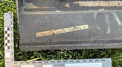 Black rectangular case on grass with a beige labeled tape and two measurement rulers nearby, suggesting field evidence tagging.