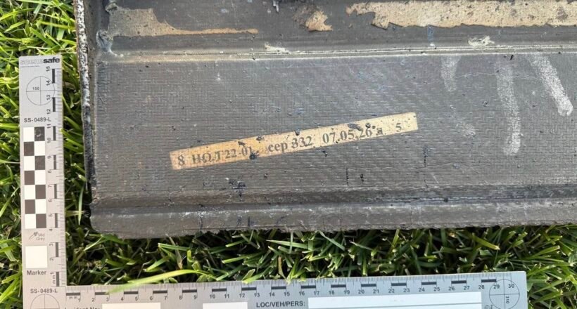 Black rectangular case on grass with a beige labeled tape and two measurement rulers nearby, suggesting field evidence tagging.