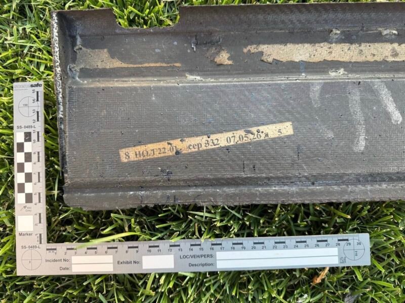 Black rectangular case on grass with a beige labeled tape and two measurement rulers nearby, suggesting field evidence tagging.