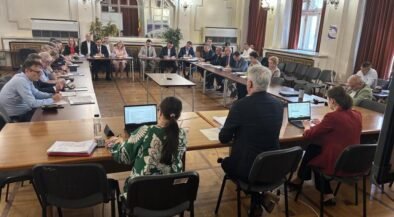 Large formal meeting in a U-shaped wooden-floored chamber with attendees seated around tables, some using laptops and papers on display.