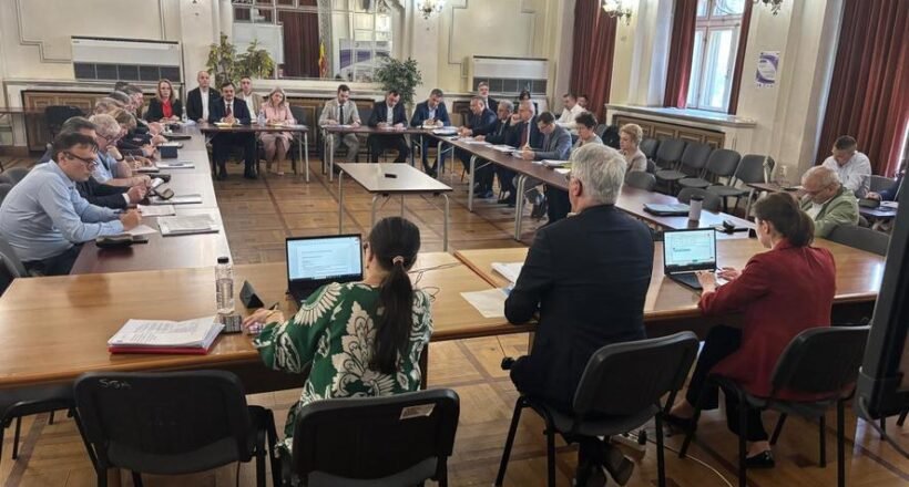 Large formal meeting in a U-shaped wooden-floored chamber with attendees seated around tables, some using laptops and papers on display.