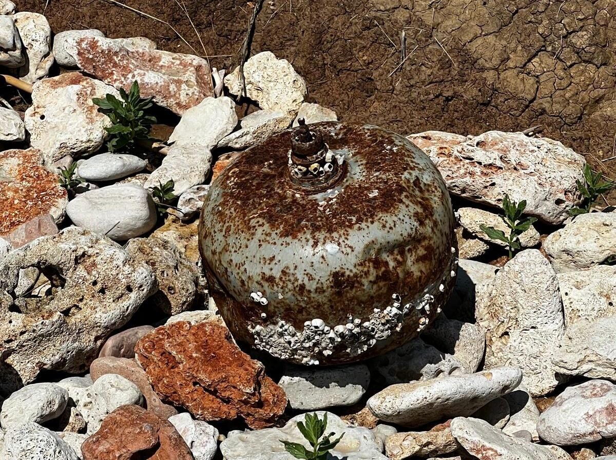 Rusty spherical buoy covered with barnacles resting among rocks on shore