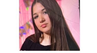 Portrait of a young woman with long dark hair wearing a black top, posed against a pink backdrop with soft makeup.