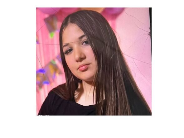 Portrait of a young woman with long dark hair wearing a black top, posed against a pink backdrop with soft makeup.