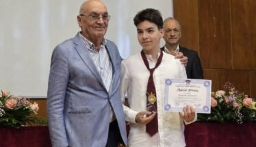 Student in a white shirt and burgundy tie holds a certificate and gold medal, flanked by two men at an awards ceremony on stage with flowers nearby.
