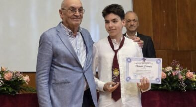 Student in a white shirt and burgundy tie holds a certificate and gold medal, flanked by two men at an awards ceremony on stage with flowers nearby.