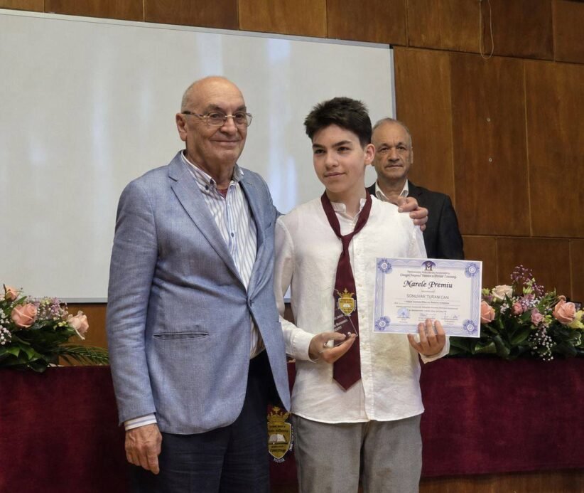 Student in a white shirt and burgundy tie holds a certificate and gold medal, flanked by two men at an awards ceremony on stage with flowers nearby.