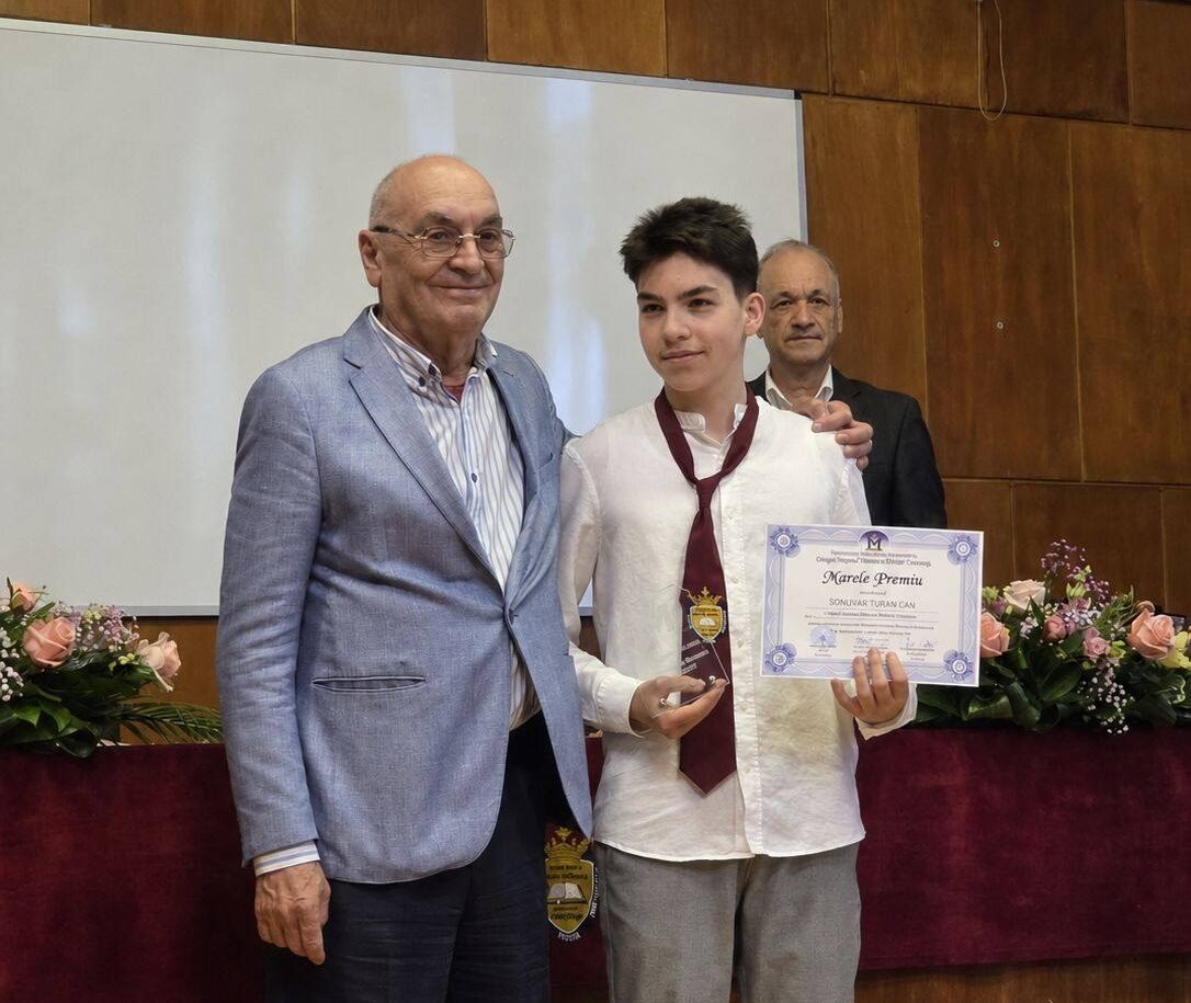 Student in a white shirt and burgundy tie holds a certificate and gold medal, flanked by two men at an awards ceremony on stage with flowers nearby.