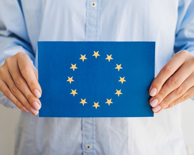 Person holding a blue card displaying the European Union flag: circle of 12 gold stars around a center space.