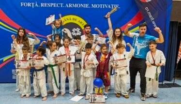 Group of children in karate gis posing with certificates and medals on a gym stage, with a coach on the right and a blue banner behind them.