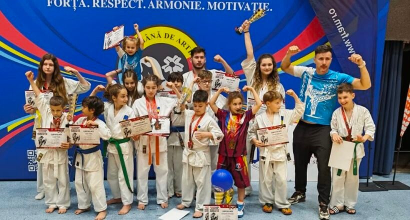 Group of children in karate gis posing with certificates and medals on a gym stage, with a coach on the right and a blue banner behind them.