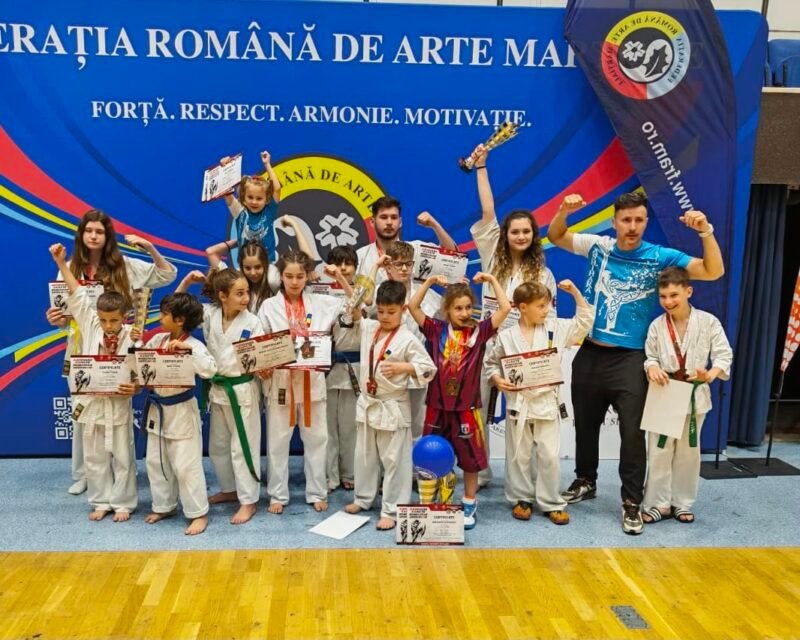 Group of children in karate gis posing with certificates and medals on a gym stage, with a coach on the right and a blue banner behind them.