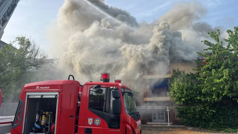 Red fire engine in the foreground as a building emits a thick plume of smoke from a fire behind it.