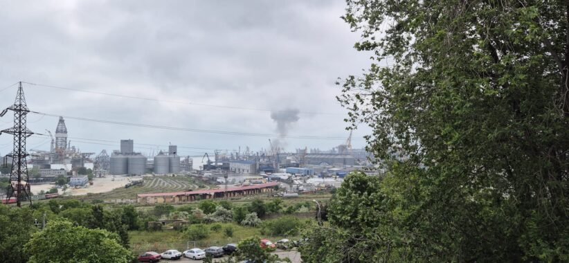 Industrial port scene with cranes, silos and a smokestack, viewed from a green hillside under a cloudy sky.