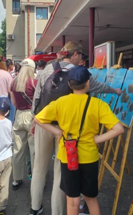 Group of people, adults and kids, reading blue exhibit panels on wooden easels outside a building.
