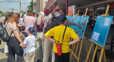 Group of people, adults and kids, reading blue exhibit panels on wooden easels outside a building.