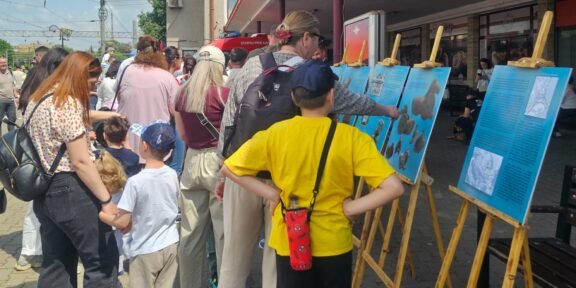 Group of people, adults and kids, reading blue exhibit panels on wooden easels outside a building.