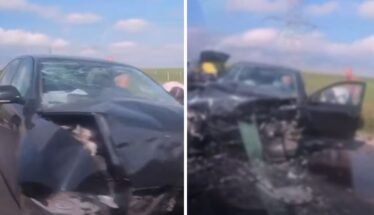 Black sedan with heavily damaged front end after a crash; airbags deployed and driver-side door open on a rural highway with debris on the road.