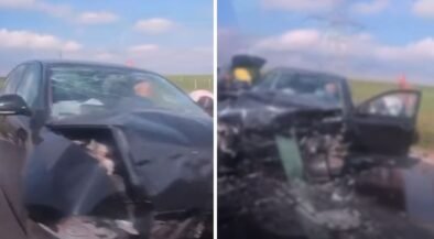 Black sedan with heavily damaged front end after a crash; airbags deployed and driver-side door open on a rural highway with debris on the road.