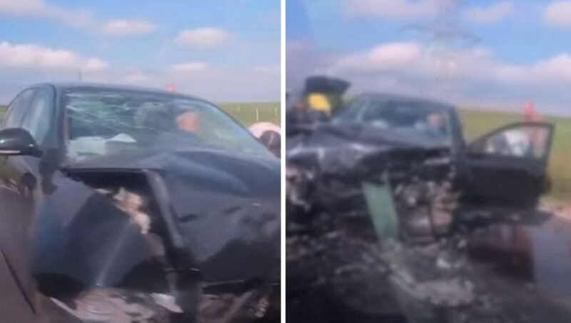 Black sedan with heavily damaged front end after a crash; airbags deployed and driver-side door open on a rural highway with debris on the road.