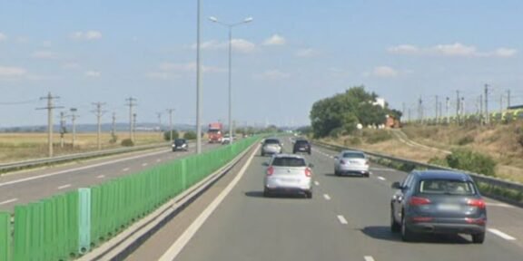 Multi-lane highway on a sunny day with green roadside barriers on the left and several cars ahead in each lane.