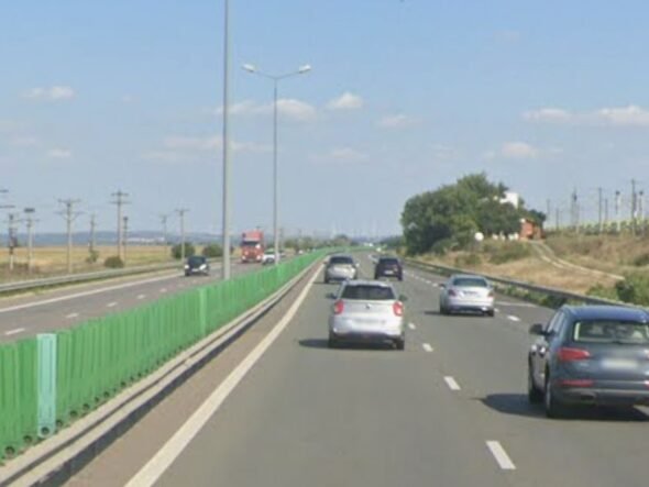 Multi-lane highway on a sunny day with green roadside barriers on the left and several cars ahead in each lane.