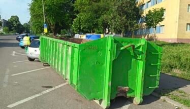 Green industrial dumpster on wheels blocking a row of parking spaces on a residential street, with trees and a yellow building in the background.