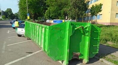 Green industrial dumpster on wheels blocking a row of parking spaces on a residential street, with trees and a yellow building in the background.