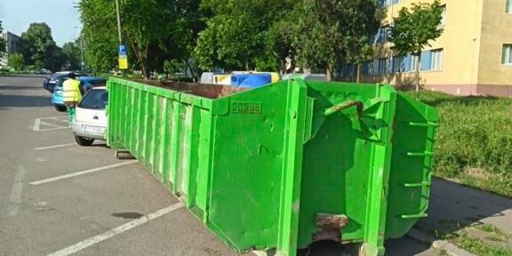 Green industrial dumpster on wheels blocking a row of parking spaces on a residential street, with trees and a yellow building in the background.