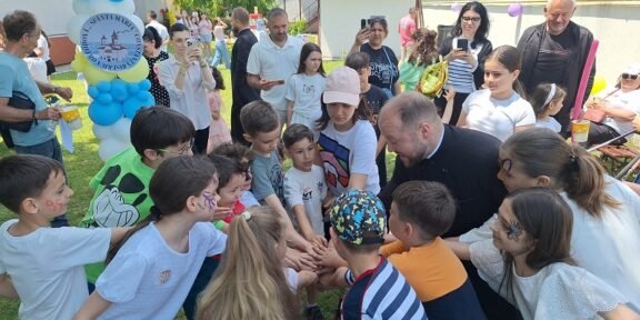 Children and adults gather in a circle with hands joined in the center at an outdoor community event, balloons in the background.