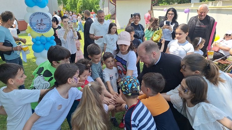Children and adults gather in a circle with hands joined in the center at an outdoor community event, balloons in the background.