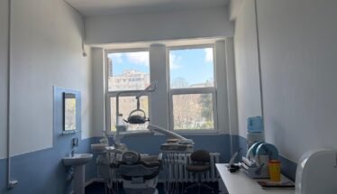 Dental treatment room with a patient chair, overhead light, and dental tools ready for a procedure near large windows.