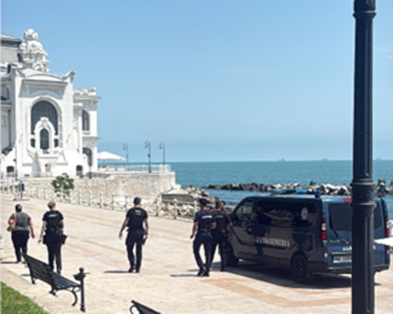 Police officers walking along a sunny seaside promenade by a white ornate building, with a dark van parked near the water.