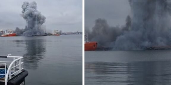 Port scene with a tall, dark smoke plume rising from a fire on the water, several ships in the distance and calm, overcast water nearby.