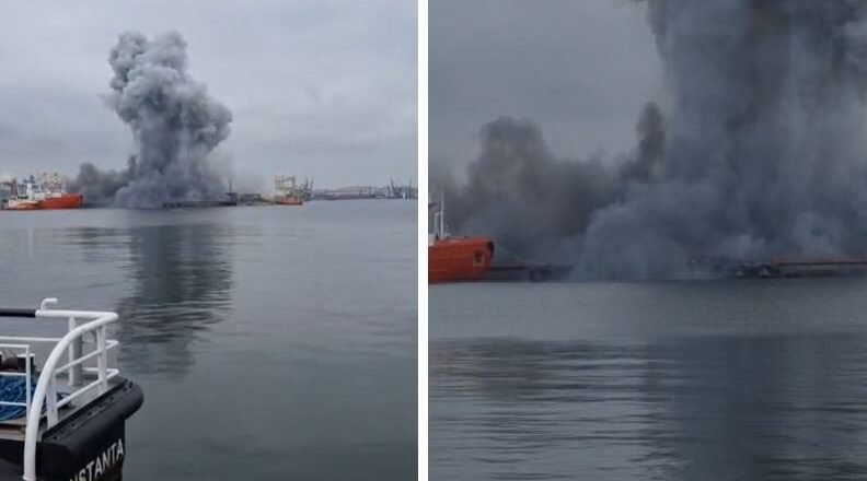 Port scene with a tall, dark smoke plume rising from a fire on the water, several ships in the distance and calm, overcast water nearby.