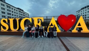 Group of people sitting on a low wall in front of large, illuminated letters spelling 'SUCEAVA' with a red heart graphic between letters.