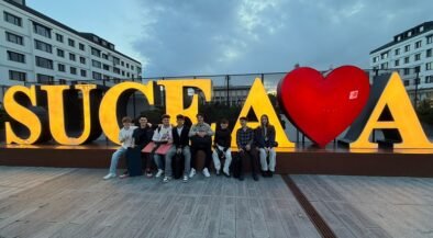Group of people sitting on a low wall in front of large, illuminated letters spelling 'SUCEAVA' with a red heart graphic between letters.