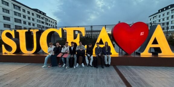 Group of people sitting on a low wall in front of large, illuminated letters spelling 'SUCEAVA' with a red heart graphic between letters.