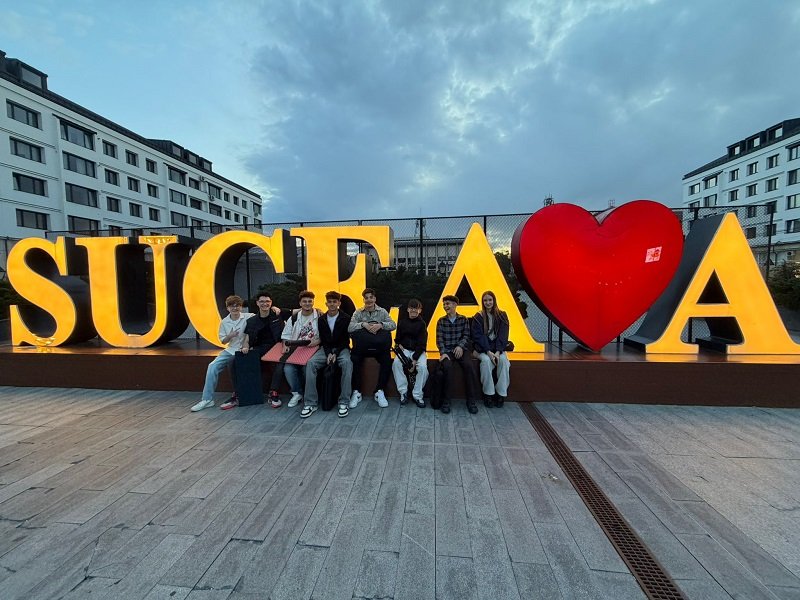 Group of people sitting on a low wall in front of large, illuminated letters spelling 'SUCEAVA' with a red heart graphic between letters.