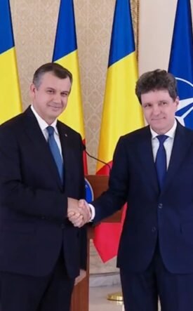 Two men in dark suits shake hands in a formal room, with EU, Romania, Ukraine, and NATO flags behind them.