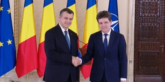 Two men in dark suits shake hands in a formal room, with EU, Romania, Ukraine, and NATO flags behind them.