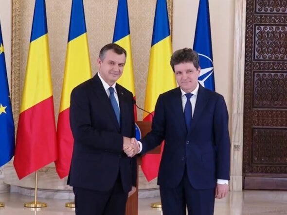 Two men in dark suits shake hands in a formal room, with EU, Romania, Ukraine, and NATO flags behind them.